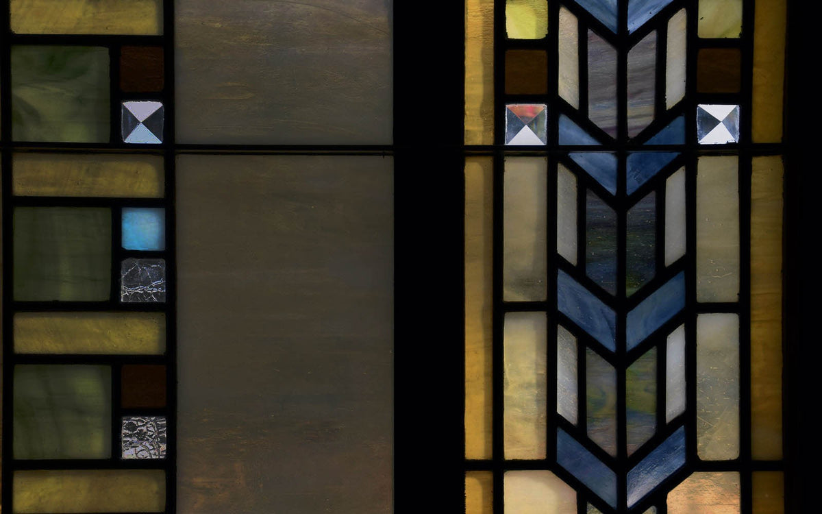 Close-up of a geometric stained-glass window, featuring blue chevron patterns and rectangular panels in muted shades of amber, green, and cream.