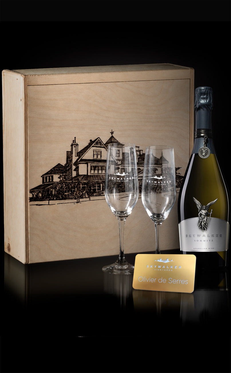 A luxury gift set featuring a wooden box engraved with the Skywalker Ranch Main House, two champagne flutes with logo, a bottle of Skywalker Sparkling wine, and an engraved gold placard on a reflective black surface.